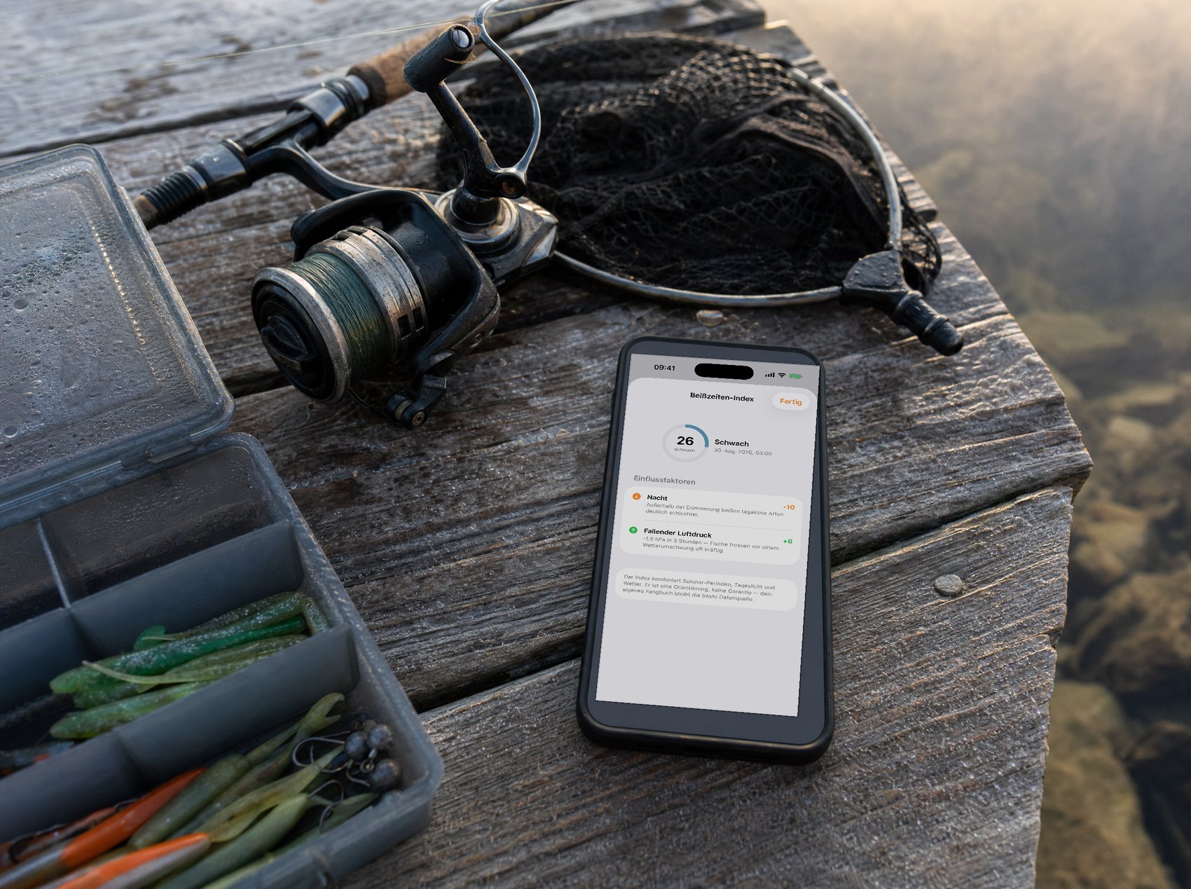 Ausrüstung auf einem Steg im Morgenlicht: Köderbox, Rolle, Kescher und ein iPhone, das die Einflussfaktoren des Beißzeiten-Index zeigt